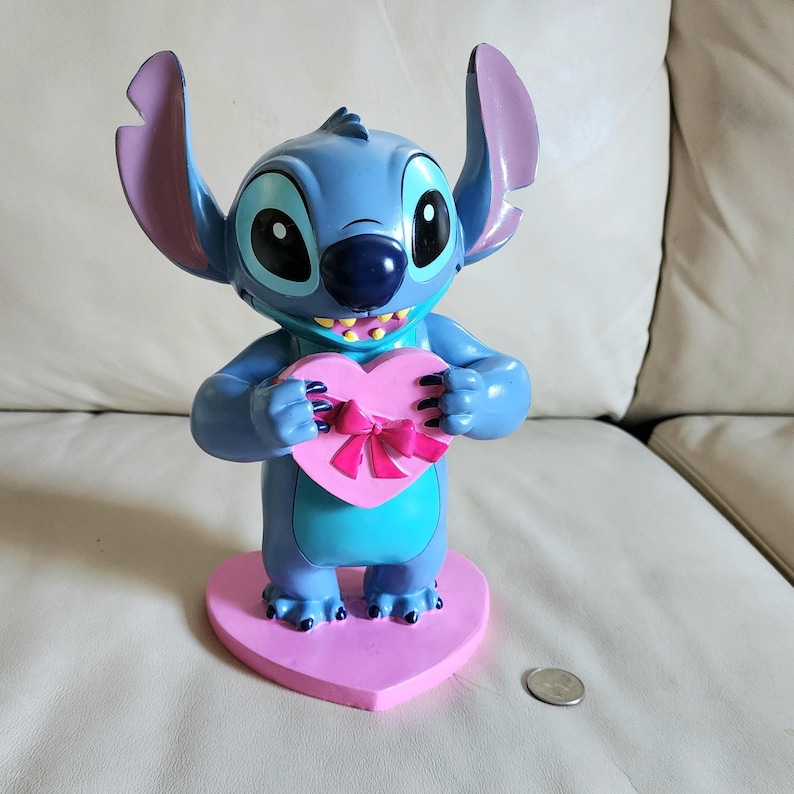 May include: A blue Stitch figurine holding a pink heart-shaped box with a pink bow. The figurine has large ears, a dark blue nose, and a wide grin. It stands on a pink heart-shaped base.