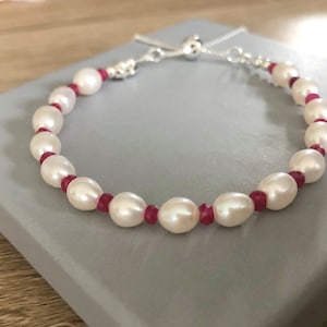 May include: A white pearl bracelet with red gemstone accents and a silver adjustable clasp.