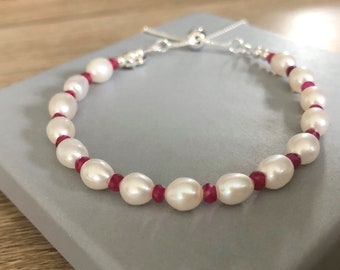 Könnte beinhalten: Ein Armband aus weißen Perlen mit roten Edelstein-Akzenten und einem verstellbaren Verschluss aus Silber.