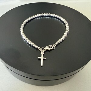 May include: A silver-toned beaded bracelet featuring a small, simple cross charm suspended from a lobster claw clasp. The bracelet is constructed from a continuous strand of uniform, polished metallic spheres that form a circular chain design.