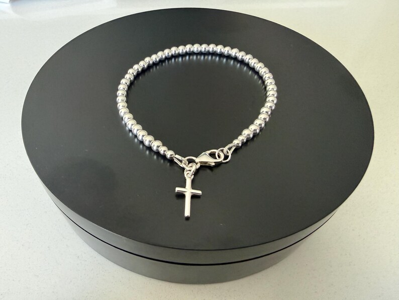 May include: A silver-toned beaded bracelet featuring a small, simple cross charm suspended from a lobster claw clasp. The bracelet is constructed from a continuous strand of uniform, polished metallic spheres that form a circular chain design.