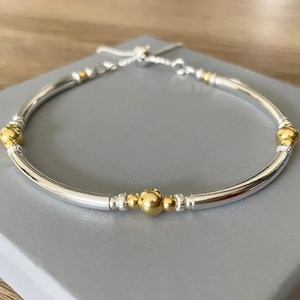 Könnte beinhalten: Ein silbernes Armband mit goldener Akzent. Das Armband ist verstellbar und hat ein schlichtes Design.