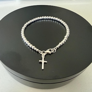 May include: A silver-coloured beaded bracelet featuring a small, simple cross charm attached to a lobster claw clasp. The bracelet is constructed from a continuous string of uniform, polished metallic spheres that form a circular band around the wrist.