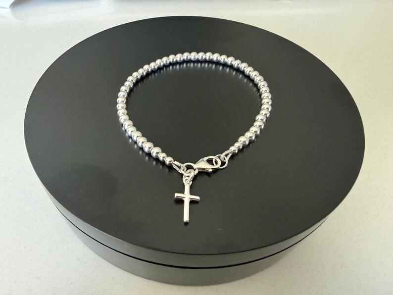 May include: A silver-coloured beaded bracelet featuring a small, simple cross charm attached to a lobster claw clasp. The bracelet is constructed from a continuous string of uniform, polished metallic spheres that form a circular band around the wrist.