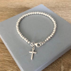 May include: A silver-toned beaded bracelet featuring a small, simple cross charm attached to a lobster claw clasp. The bracelet is constructed from a continuous strand of uniform, polished metallic spheres that form a circular band around the wrist.