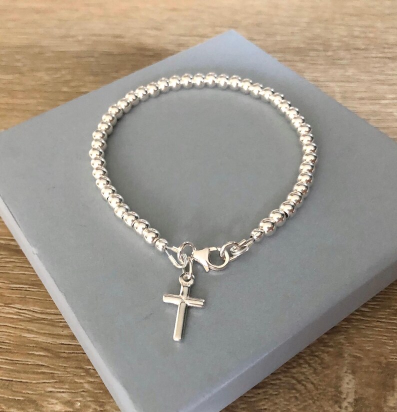 May include: A silver-toned beaded bracelet featuring a small, simple cross charm attached to a lobster claw clasp. The bracelet is constructed from a continuous strand of uniform, polished metallic spheres that form a circular band around the wrist.