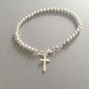 May include: A silver-coloured beaded bracelet featuring a series of small, uniform spherical beads strung together. The bracelet includes a lobster claw clasp and a small, simple silver-coloured cross charm hanging from the centre of the closure mechanism.
