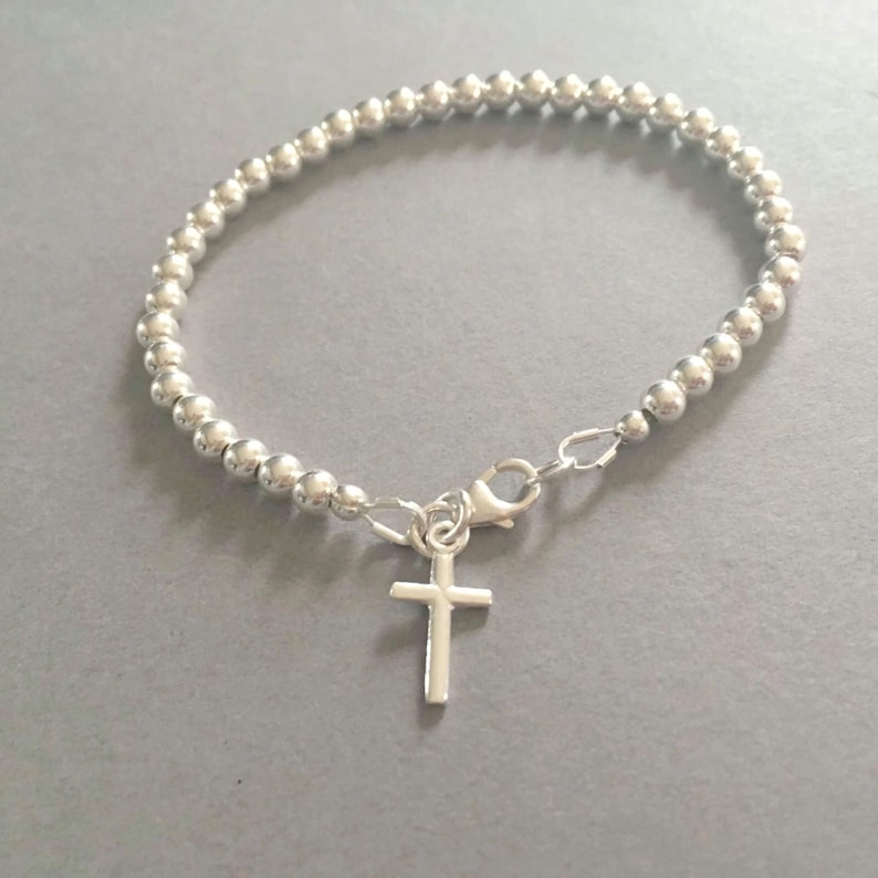May include: A silver-coloured beaded bracelet featuring a series of small, uniform spherical beads strung together. The bracelet includes a lobster claw clasp and a small, simple silver-coloured cross charm hanging from the centre of the closure mechanism.