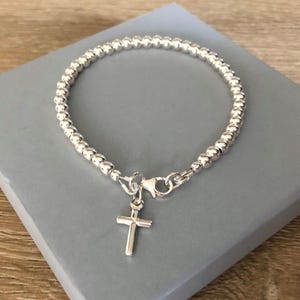 May include: A silver-toned beaded bracelet featuring a small, polished cross charm suspended from a lobster claw clasp. The bracelet is constructed from a continuous strand of uniform, reflective spherical beads arranged in a circular shape.