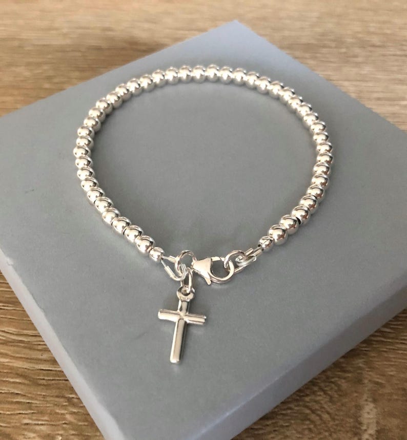 May include: A silver-toned beaded bracelet featuring a small, polished cross charm suspended from a lobster claw clasp. The bracelet is constructed from a continuous strand of uniform, reflective spherical beads arranged in a circular shape.