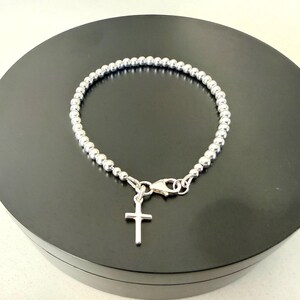 May include: A silver-toned beaded bracelet featuring a small, plain cross charm suspended from a lobster claw clasp. The bracelet is constructed from a continuous strand of uniform, polished spherical beads that form a circular shape.