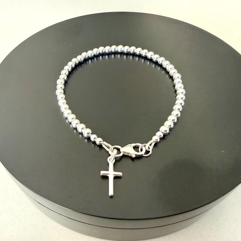 May include: A silver-toned beaded bracelet featuring a small, plain cross charm suspended from a lobster claw clasp. The bracelet is constructed from a continuous strand of uniform, polished spherical beads that form a circular shape.