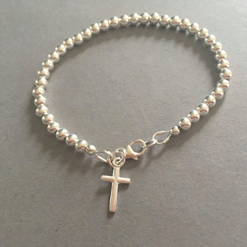 May include: A silver-toned beaded bracelet featuring a series of small, uniform spherical beads strung together. The bracelet includes a lobster claw clasp and a small, simple cross charm hanging from the closure mechanism.