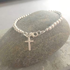 May include: A silver-toned beaded bracelet featuring a small, plain cross charm suspended from a lobster claw clasp. The bracelet is constructed from a continuous strand of uniform, polished metallic spheres that form a circular band around the wrist.