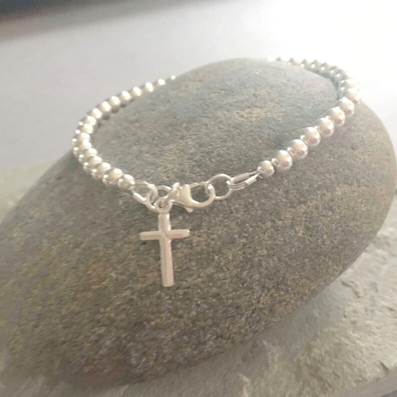May include: A silver-toned beaded bracelet featuring a small, plain cross charm suspended from a lobster claw clasp. The bracelet is constructed from a continuous strand of uniform, polished metallic spheres that form a circular band around the wrist.