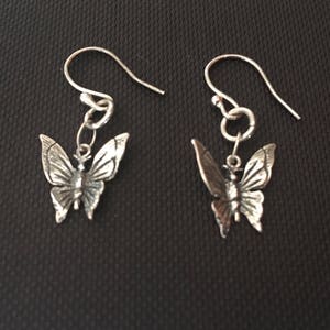 K&ouml;nnte beinhalten: Ein Paar silberne Schmetterlingsohrringe. Jeder Ohrring hat einen detaillierten Schmetterlingsanh&auml;nger, der an einem einfachen Haken h&auml;ngt. Die Schmetterlingsfl&uuml;gel sind aufwendig gestaltet und weisen eine zarte, strukturierte Oberfl&auml;che auf. Die Ohrringe sind vor dunklem Hintergrund.
