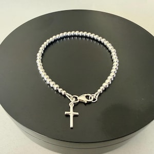 May include: A silver-toned beaded bracelet featuring a small, plain cross charm hanging from a lobster claw clasp. The bracelet is constructed from a continuous strand of uniform, polished metallic spheres that form a circular chain design.