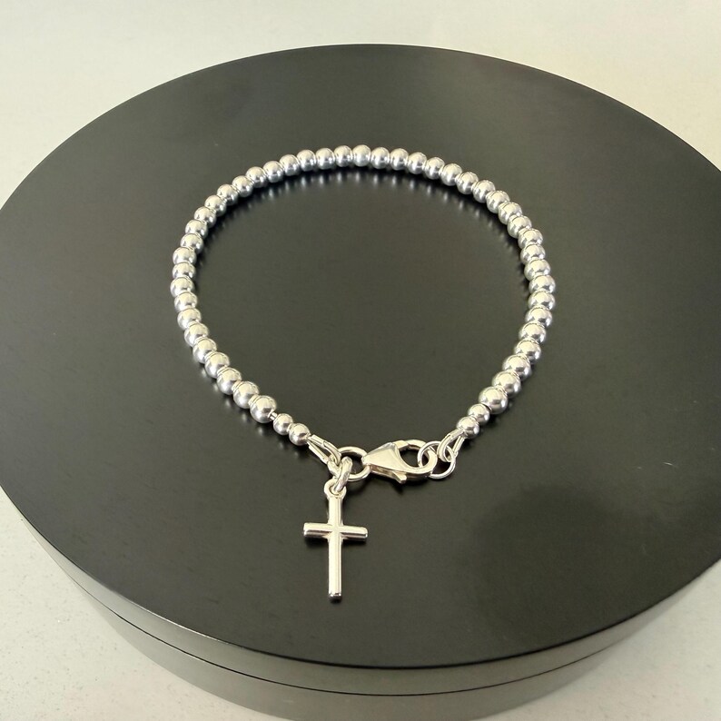 May include: A silver-toned beaded bracelet featuring a small, plain cross charm hanging from a lobster claw clasp. The bracelet is constructed from a continuous strand of uniform, polished metallic spheres that form a circular chain design.