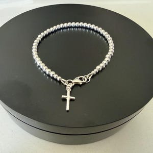 May include: A silver-coloured beaded bracelet featuring a small, simple cross charm suspended from a lobster claw clasp. The bracelet is constructed from a continuous strand of uniform, polished metallic spheres that form a circular chain link design.