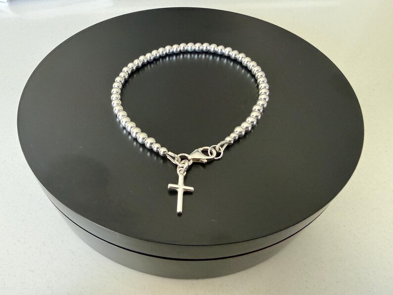 May include: A silver-coloured beaded bracelet featuring a small, simple cross charm suspended from a lobster claw clasp. The bracelet is constructed from a continuous strand of uniform, polished metallic spheres that form a circular chain link design.