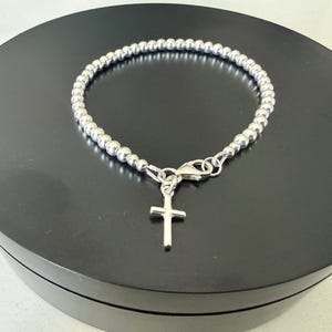 May include: A silver-toned beaded bracelet featuring a small, plain cross charm suspended from a lobster claw clasp. The bracelet is constructed from a continuous strand of uniform, polished spherical beads that form a circular shape.