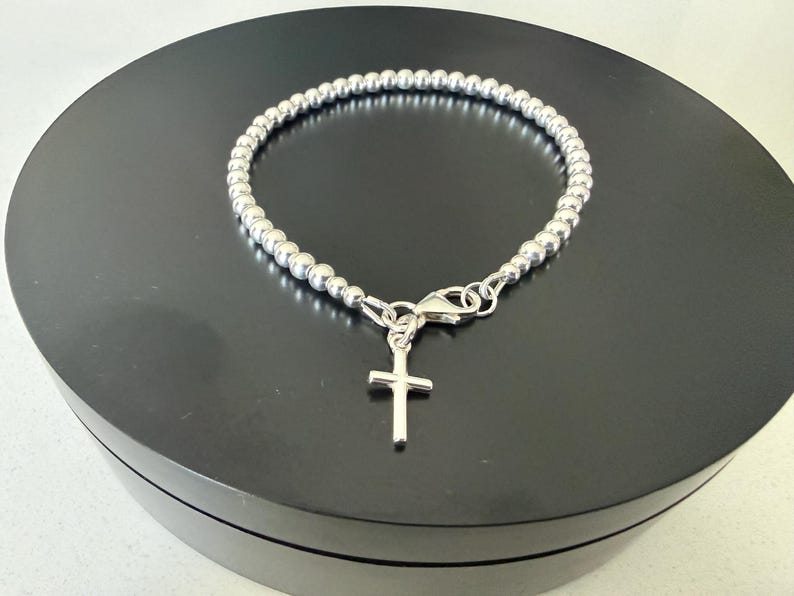 May include: A silver-toned beaded bracelet featuring a small, plain cross charm suspended from a lobster claw clasp. The bracelet is constructed from a continuous strand of uniform, polished spherical beads that form a circular shape.
