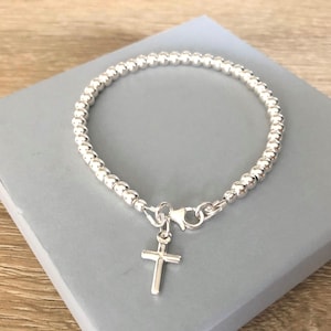 May include: A silver-toned beaded bracelet featuring a small, simple cross charm suspended from a lobster claw clasp. The bracelet is constructed from a continuous strand of uniform, polished spherical beads that create a circular shape.