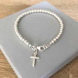 May include: A silver-toned beaded bracelet featuring a small, simple cross charm attached to a lobster claw clasp. The bracelet is constructed from a continuous strand of uniform, polished spherical beads that form a circular shape.