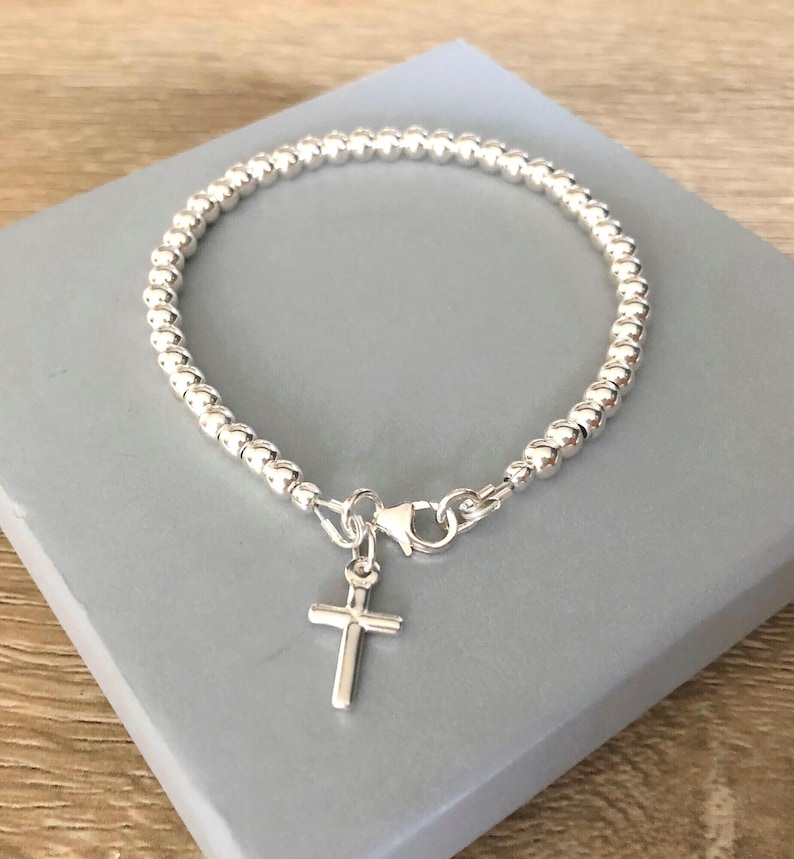 May include: A silver-toned beaded bracelet featuring a small, simple cross charm attached to a lobster claw clasp. The bracelet is constructed from a continuous strand of uniform, polished spherical beads that form a circular shape.