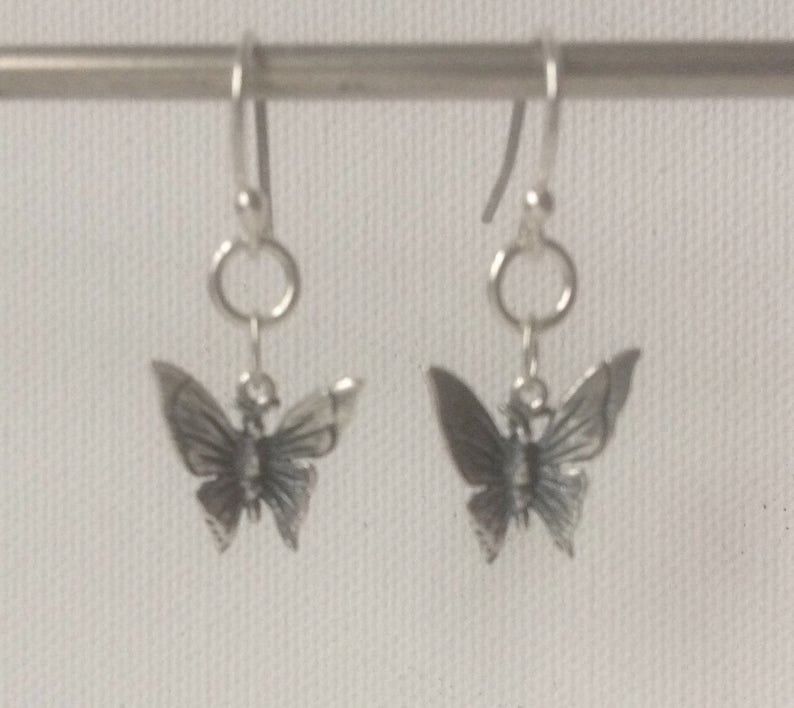 K&ouml;nnte beinhalten: Ein Paar silberfarbene Schmetterlingsohrringe. Jeder Ohrring hat einen detaillierten Schmetterlingsanh&auml;nger, der an einem Haken h&auml;ngt. Die Schmetterlingsfl&uuml;gel sind ausgebreitet und das Design ist aufwendig. Die Ohrringe werden vor einem schlichten Hintergrund gezeigt.