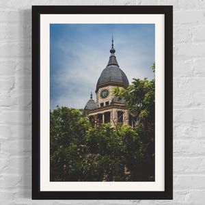 May include: A black framed photograph of a stone building with a clock tower. The building is partially obscured by green trees. The sky is blue with white clouds.