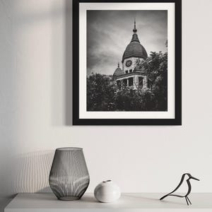 May include: Black and white photograph of a stone building with a clock tower. The building is surrounded by trees and the sky is cloudy.