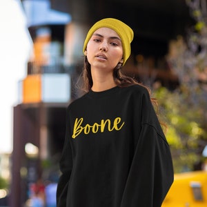 以下が含まれることがあります： 黄色の筆記体で「Boone」と書かれた黒いスウェットシャツ。モデルは黄色のビーニーを着用している。