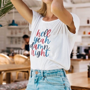 Op de afbeelding: Een wit T-shirt met de tekst "hell yeah damn right" in rode, blauwe en witte letters.
