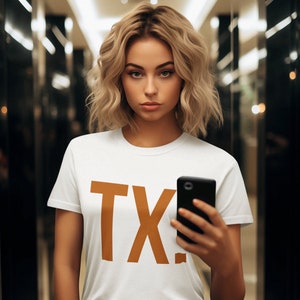 May include: A woman wearing a white t-shirt with the text "TX" printed in brown on the front. She is holding a black smartphone in her right hand.