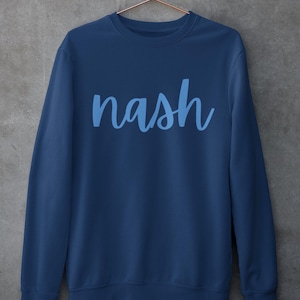 以下が含まれることがあります： 前面に薄い青色の「nash」という文字がプリントされた紺色のスウェットシャツです。