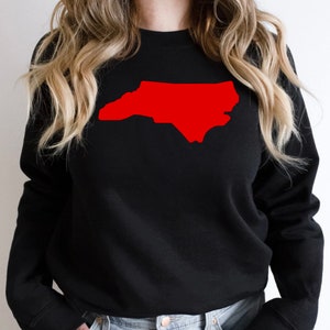 May include: Black sweatshirt featuring a bold red outline of the state of North Carolina. The long-sleeved top is made of a soft material, perfect for casual wear. A simple, yet striking design for those who love North Carolina.