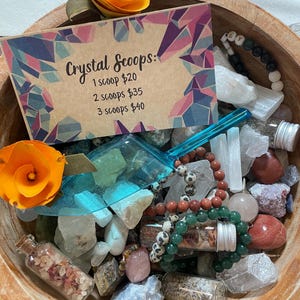 May include: A wooden bowl filled with various crystals, gemstones, and small glass vials. The bowl contains a variety of colors and textures, including clear, pink, and brown stones. A card reads "Crystal Scoops". Two orange paper flowers are placed on the rim.