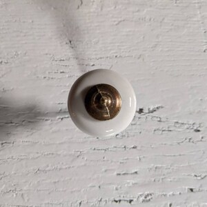 Op de afbeelding: Witte keramische knop met een gouden middenstuk. De knop bevindt zich op een wit houten oppervlak.