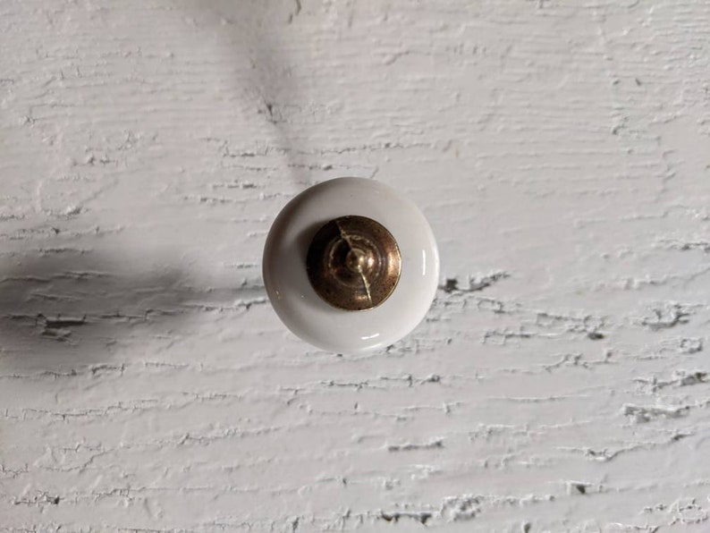 Op de afbeelding: Witte keramische knop met een gouden middenstuk. De knop bevindt zich op een wit houten oppervlak.