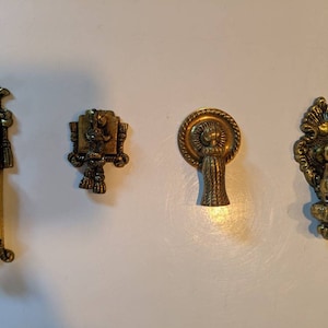 May include: Four antique brass cabinet knobs with ornate designs. The knobs have different shapes and styles, including a pull with a tassel, a square knob with a raised design, and two round knobs with intricate details.