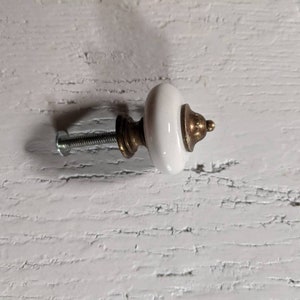 Op de afbeelding: Witte keramische knop met een gouden metalen basis en een schroef. De knop is bevestigd aan een wit houten oppervlak.