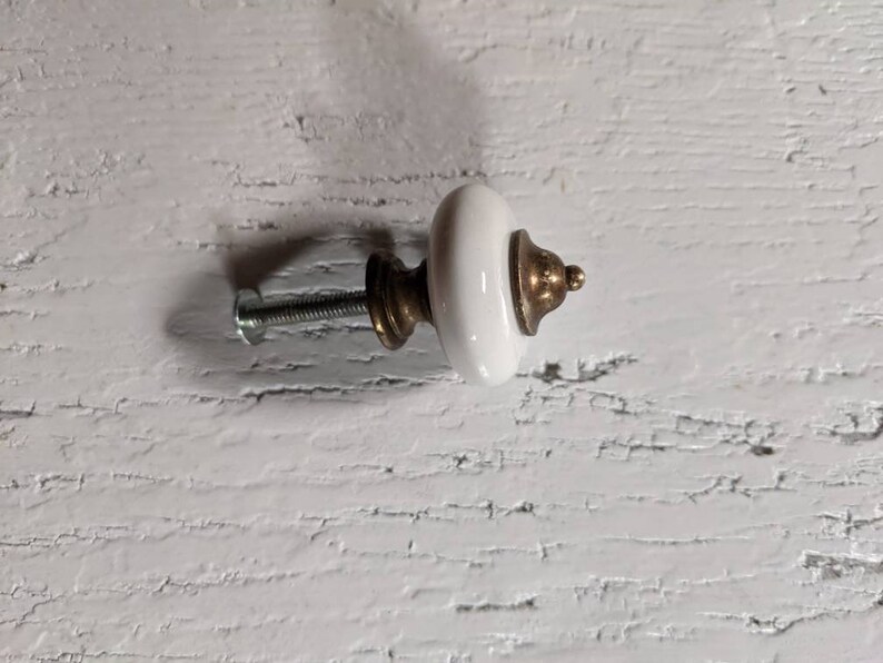 Op de afbeelding: Witte keramische knop met een gouden metalen basis en een schroef. De knop is bevestigd aan een wit houten oppervlak.