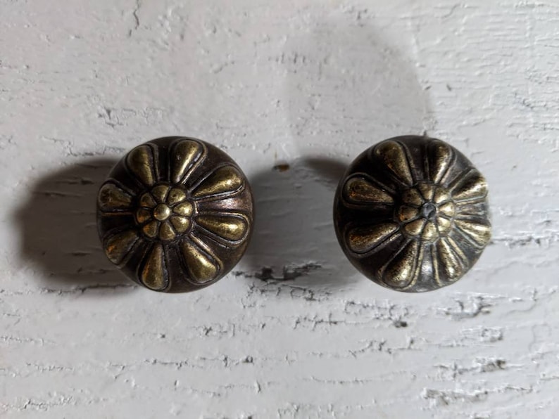 Op de afbeelding: Twee ronde, decoratieve knoppen met een messing afwerking. De knoppen hebben een reli&euml;f, bloemmotief met een centrale bloem.