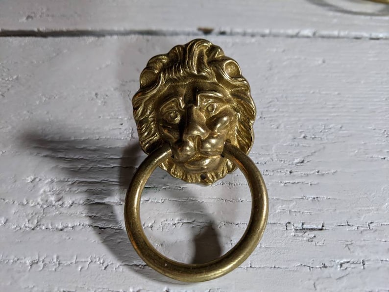 Op de afbeelding: Een gouden deurknop met een leeuwenkop en een ronde ring.