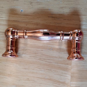 May include: A rose gold cabinet pull with a rounded, slightly curved bar and rounded ends. The pull is made of metal and has a polished finish.