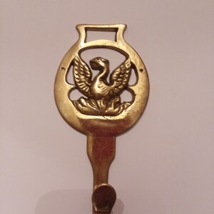 May include: A gold-toned metal hook with a decorative design featuring a bird in an oval shape. The hook is attached to a wall.