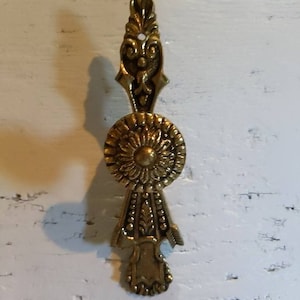 May include: A gold-toned metal drawer pull with an ornate design. The pull features a circular center with a floral pattern and a pointed top with intricate details.