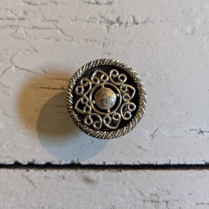 Op de afbeelding: Een zilveren decoratieve knop met een cirkelvormig ontwerp met een centrale ronde knop en ingewikkelde krulwerk.