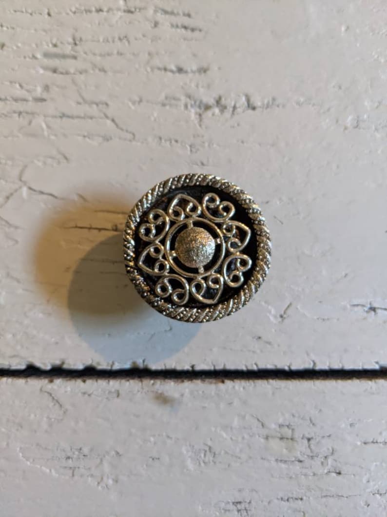 Op de afbeelding: Een zilveren decoratieve knop met een cirkelvormig ontwerp met een centrale ronde knop en ingewikkelde krulwerk.